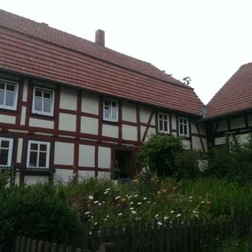 L'image représente une maison traditionnelle de style allemand, avec un toit en tuiles rouges et des murs blancs, entourée d'une végétation luxuriante et d'une clôture en bois au premier plan.