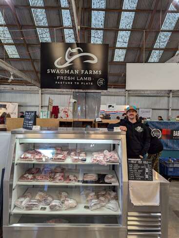 Ein Mann steht hinter einer Fleischauslage bei Swagman Farms und verkauft frische Lammfleischstücke. Der Stand ist mit Schildern, Preislisten und verpackten Lammfleischprodukten ausgestattet.