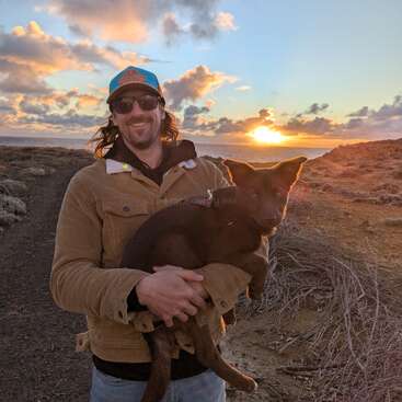 Un homme portant des lunettes de soleil et un chapeau sourit en tenant un chien brun. Ils sont en plein air sur un chemin rocailleux avec un magnifique coucher de soleil et des nuages derrière eux.