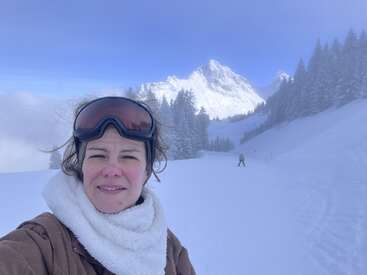 Une femme en tenue de ski sourit pour un selfie sur une pente enneigée, entourée de pins, avec en arrière-plan un majestueux sommet recouvert de neige.
