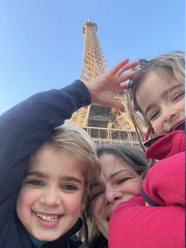Un selfie de famille plein de joie pris au pied de la Tour Eiffel, sur lequel on voit deux enfants souriants et un adulte, tous profitant d'une belle journée ensoleillée.