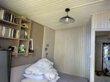 Una habitación pequeña y acogedora con paredes y techo de madera clara, estanterías repletas de libros, una cama con la ropa de cama bien doblada, una lámpara original y un perro a su lado.