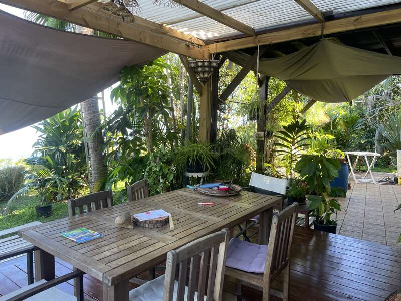 Un acogedor patio al aire libre cuenta con una mesa y sillas de madera rodeadas de exuberantes plantas tropicales, a la sombra de lonas y una pérgola de madera, lo que crea un ambiente relajante.