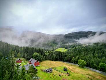 Une vallée brumeuse et luxuriante avec des forêts vertes, des collines ondulantes et des maisons rouges éparpillées. Le ciel couvert ajoute une atmosphère sereine et paisible à ce paysage rural.