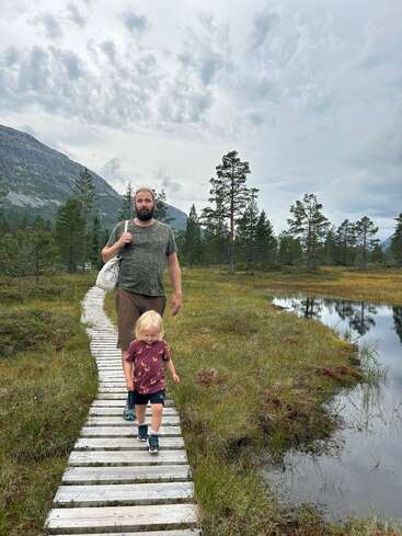 Un homme et un jeune enfant marchent sur un chemin de bois dans une zone humide pittoresque et herbeuse, avec des arbres, des montagnes et un ciel nuageux en arrière-plan.
