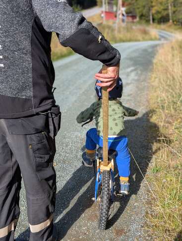 Un enfant apprend à faire du vélo sur une route de gravier, guidé par un adulte qui tient une poignée en bois attachée au vélo pour le soutenir.