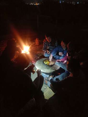 Um grupo de pessoas senta ao redor de uma fogueira à noite, compartilhando comida em uma pequena mesa, curtindo o calor e a companhia sob o céu escuro e estrelado.