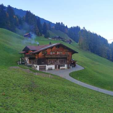 A charming wooden chalet sits on a lush green hillside, surrounded by forested mountains. Smoke rises from the chimney, and other houses dot the scenic landscape.