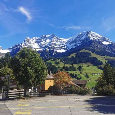 Snow-capped mountains rise majestically above a charming village with green hills, scattered houses, and colorful trees under a clear blue sky, creating a picturesque landscape.