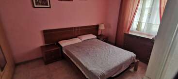 This cozy bedroom features a double bed with a striped cover, two bedside tables, a lamp, dresser, artwork on pink walls, and a window with sheer curtains.