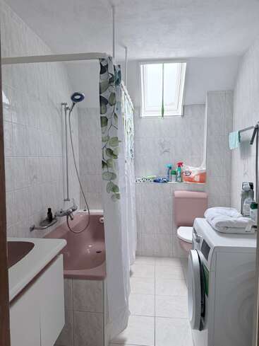 This small bathroom features a pink bathtub, toilet, white vanity, and washing machine. A shower curtain, cleaning supplies, towels, and a skylight complete the room.