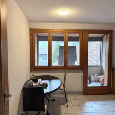 A cozy room features a round table with two chairs by large wooden-framed windows, letting in natural light. Outside, a balcony holds shopping bags and toys.