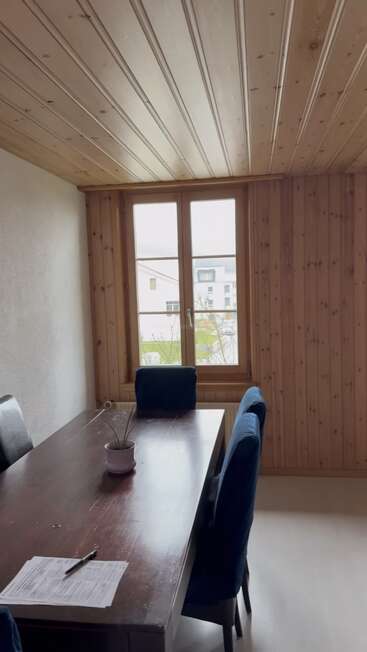 A cozy room with wooden walls and ceiling, a dark dining table, blue chairs, a small potted plant, and paperwork on the table near a window.