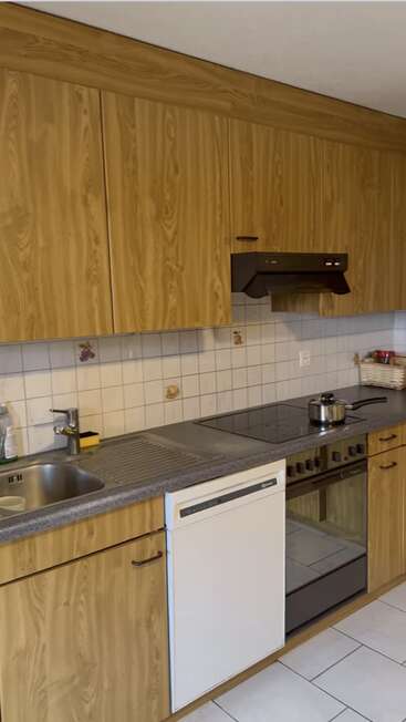 This image shows a kitchen with wooden cabinets, white tiled backsplash, sink, dishwasher, electric stovetop, oven, pots, soap, and a basket containing some jars.