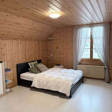 This cozy bedroom features wooden walls and ceiling, a comfortable bed with pillows and a white comforter, a window with sheer curtains, and minimalistic decor.
