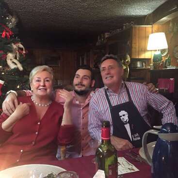 L'image représente une scène festive où trois personnes posent pour une photo à une table, entourées de décorations de Noël et d'une bouteille de vin.