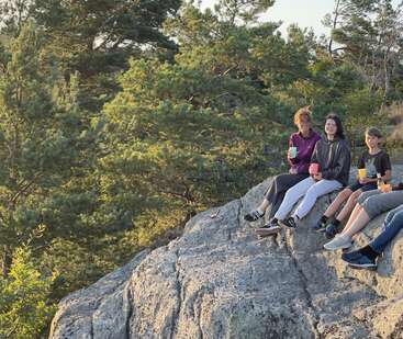 The image depicts three individuals sitting on a rocky outcrop, surrounded by trees, with two of them holding drinks in colorful cups.