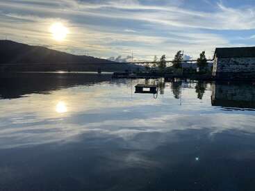 A imagem mostra um lago tranquilo ao pôr do sol, com reflexos do sol e das nuvens na água. No fundo, há uma ponte e alguns prédios.