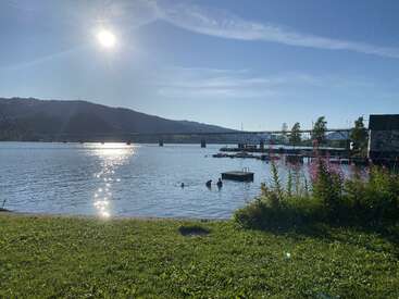 A imagem mostra um dia ensolarado e claro à beira de um lago tranquilo, com pessoas nadando, um cais, uma margem gramada, flores, uma ponte e colinas ao redor.