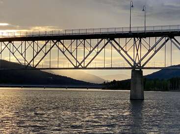 Uma ponte de aço atravessa um rio tranquilo ao pôr do sol, recortada contra a luz suave. As montanhas se erguem ao fundo, enquanto nuvens e tons dourados preenchem o céu.