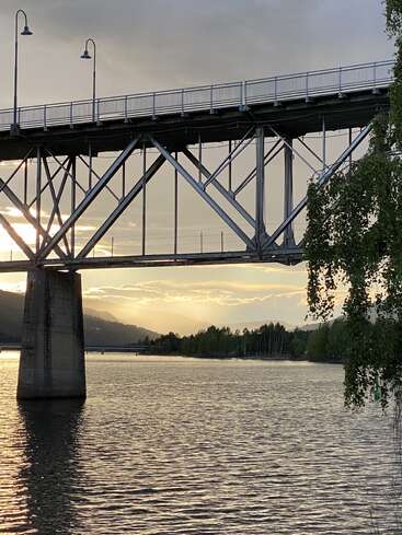 Uma ponte atravessa um rio calmo ao pôr do sol, com a luz suave do sol brilhando através dela. As árvores e as montanhas distantes emolduram lindamente a paisagem tranquila e pitoresca.