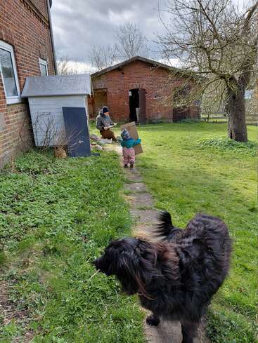 Ein großer schwarzer Hund steht auf einem grasbewachsenen Weg. Ein Kleinkind trägt einen Karton auf einen Erwachsenen zu, der in der Nähe eines Schuppens sitzt. Sie sind von Backsteinhäusern und Bäumen umgeben.