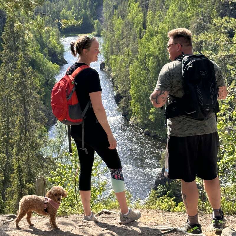 The image depicts a serene scene of a man and woman standing on a cliff overlooking a river, accompanied by a small dog, surrounded by lush trees and greenery.