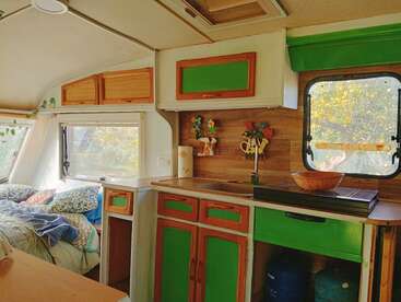 O interior desse campista aconchegante apresenta armários em tons de verde, uma pequena área de cozinha, uma cama com cobertores coloridos, ganchos decorativos na parede e grandes janelas que deixam entrar a luz do sol.