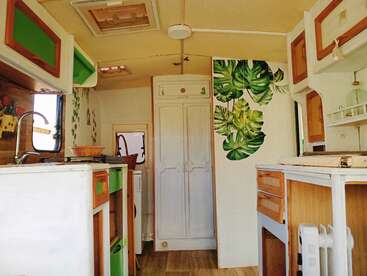 Esta imagem mostra o interior de um campista aconchegante com armários brancos e de madeira, detalhes em verde, arte na parede com folhas, uma pequena cozinha e uma atmosfera caseira e brilhante.