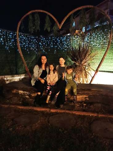 Quatro pessoas, duas mulheres e duas crianças, sentam-se juntas em um tronco à noite, sob uma grande moldura em forma de coração e luzes de fadas, desfrutando de um momento de alegria.