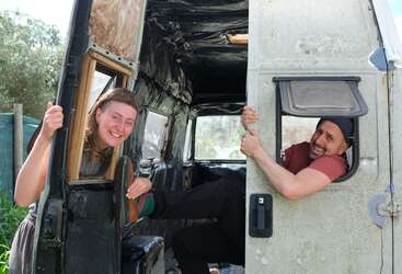 A woman and man playfully pose in a partially restored van. She smiles through the door, while he laughs, half-emerging from another opening, both joyful.