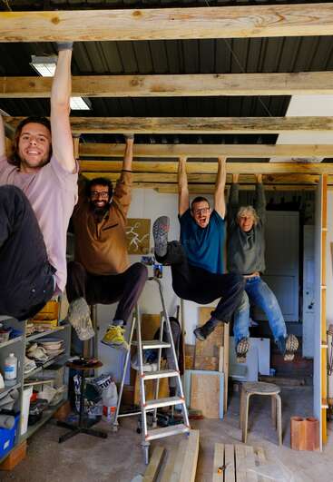 Four smiling people hang playfully from wooden beams in a workshop, with tools, shelves, and ladders visible. They appear energetic, joyful, and ready for fun.