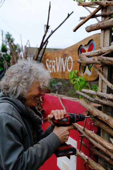 A smiling person with curly gray hair uses a power drill to construct or repair a wooden fence, beside a colorful "SERVIVO" sign and greenery.
