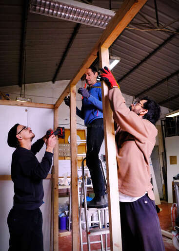 Three people are building a wooden structure indoors. One stands on a ladder, others assist by holding wood and using tools. They work together in a workshop space.