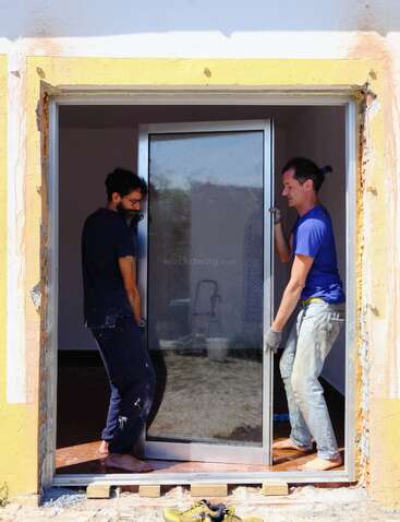 Two men are carefully installing a large glass door in a house. Both are barefoot, focused, and working together on a bright, sunny day.