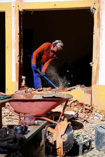 A person in safety gear breaks down a brick wall with a sledgehammer, surrounded by construction tools, debris, a wheelbarrow, and a circular saw.
