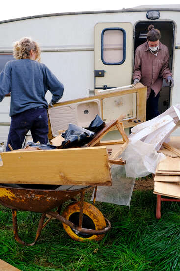 Two people dismantle and remove old furniture and debris from a caravan, loading broken parts into a rusty wheelbarrow, working outdoors on green grass.