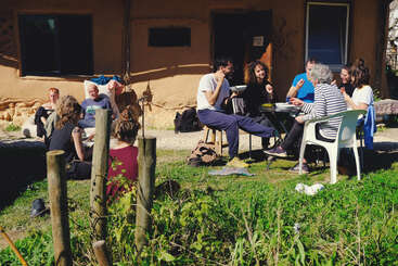 A group of people enjoy a meal together outdoors, sitting on chairs and the grass, near a rustic house, surrounded by greenery and sunlight.