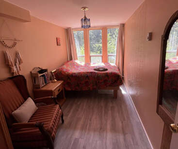 Une chambre à coucher confortable avec de grandes fenêtres, une lumière naturelle vive, un couvre-lit coloré, une chaise vintage, une petite bibliothèque, une table de nuit, des planchers en bois et des œuvres d'art murales décoratives.