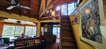 Das Bild zeigt ein geräumiges Wohnzimmer mit einem Loft, in dem ein großes Gemälde, eine Treppe und ein Esstisch zu sehen sind, umgeben von Fenstern und einer Holzdecke.