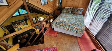 Das Bild zeigt ein gemütliches Zimmer mit einem Bett, Holzwänden und einer Treppe, die zu einem anderen Raum führt, mit einem roten Teppich und einem Fenster mit Jalousien.