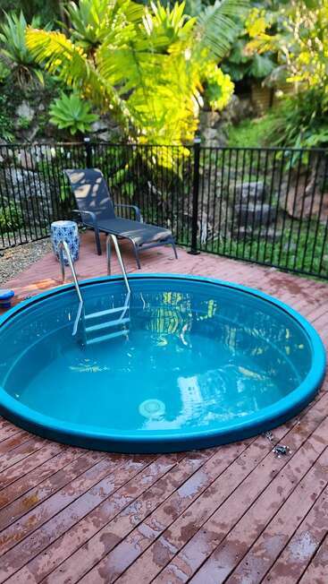 Das Bild zeigt einen kleinen, runden Pool auf einer Terrasse, umgeben von einem schwarzen Zaun, mit einem Stuhl und einer Pflanze in der Nähe, vor einer üppigen grünen Kulisse.