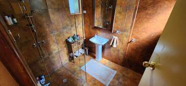 Das Bild zeigt ein Badezimmer mit Dusche, Waschbecken und Spiegel, mit braunen Fliesen an den Wänden und auf dem Boden, mit einem weißen Teppich und einem Türgriff.