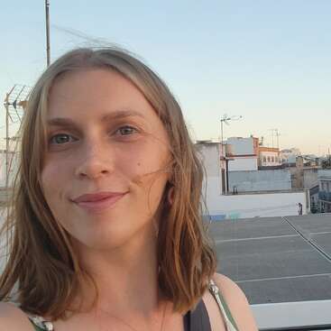 Eine Frau mit hellbraunem Haar lächelt in die Kamera. Sie steht unter freiem Himmel auf einem Dach, hinter dem Gebäude und Antennen zu sehen sind.