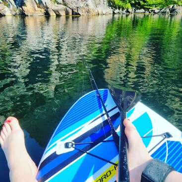 A person sits on a blue and white paddleboard with a paddle and fishing rod, enjoying calm, reflective water near rocky, tree-lined shore on a sunny day.