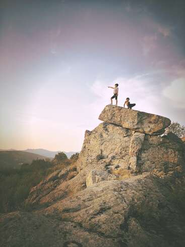 Zwei Menschen stehen auf einer großen Felsformation, der eine zeigt, der andere sitzt, inmitten einer Berglandschaft unter einem bewölkten Himmel.