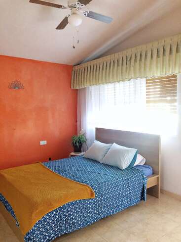 Das Bild zeigt ein Schlafzimmer mit einem Bett, einem Deckenventilator und einem Fenster, mit einer blau gemusterten Bettdecke, weißen Kissen und einer gelben Decke vor einer orangefarbenen Wand.