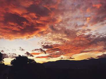 Das Bild zeigt einen atemberaubenden Sonnenuntergang über den Bergen mit leuchtend orangefarbenen und rosafarbenen Wolken, silhouettierten Bäumen und einer ruhigen Landschaft, die ein Gefühl von natürlicher Schönheit und Stille hervorruft.