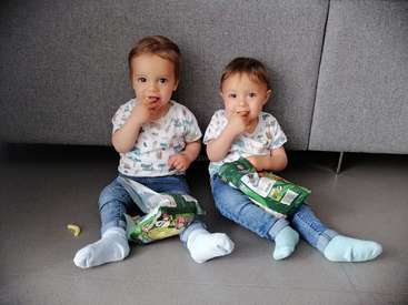 Deux enfants en bas âge sont assis par terre, chacun tenant un sac de friandises, portant des vêtements assortis, l'un mangeant une friandise et l'autre portant une friandise à sa bouche.