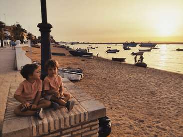 Deux jeunes garçons sont assis sur un banc de pierre, surplombant une plage de sable avec des bateaux et des eaux calmes, dans un ciel chaud de coucher de soleil.